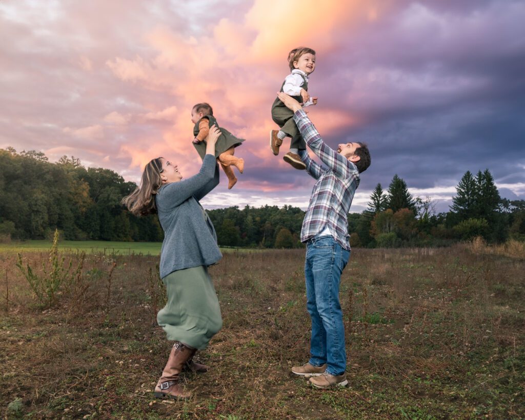 Parents lifting two young children into the air at sunset during a joyful family photo session in a wide open field near Boston.