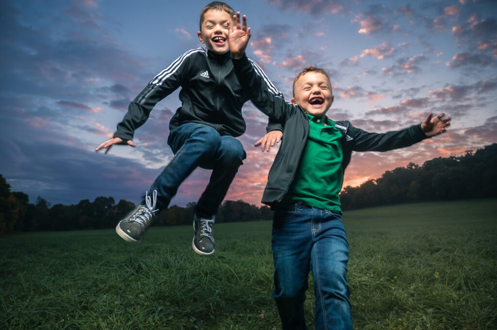 Two young brothers laughing and jumping midair in a grassy field at sunset, captured in a playful Boston-area family photo session.