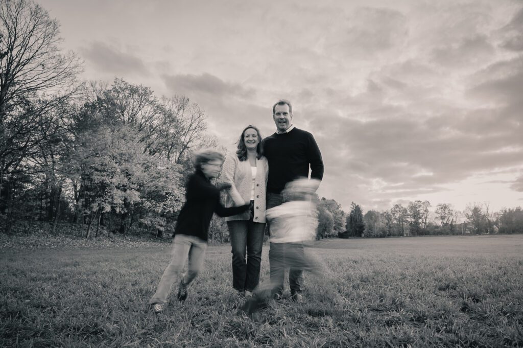 Parents smiling while their two kids blur past in motion during a black and white photo session in an open field with dramatic sky.