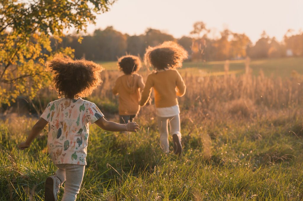 Three young children running through tall grass at golden hour during a carefree, sun-drenched family session near Boston.