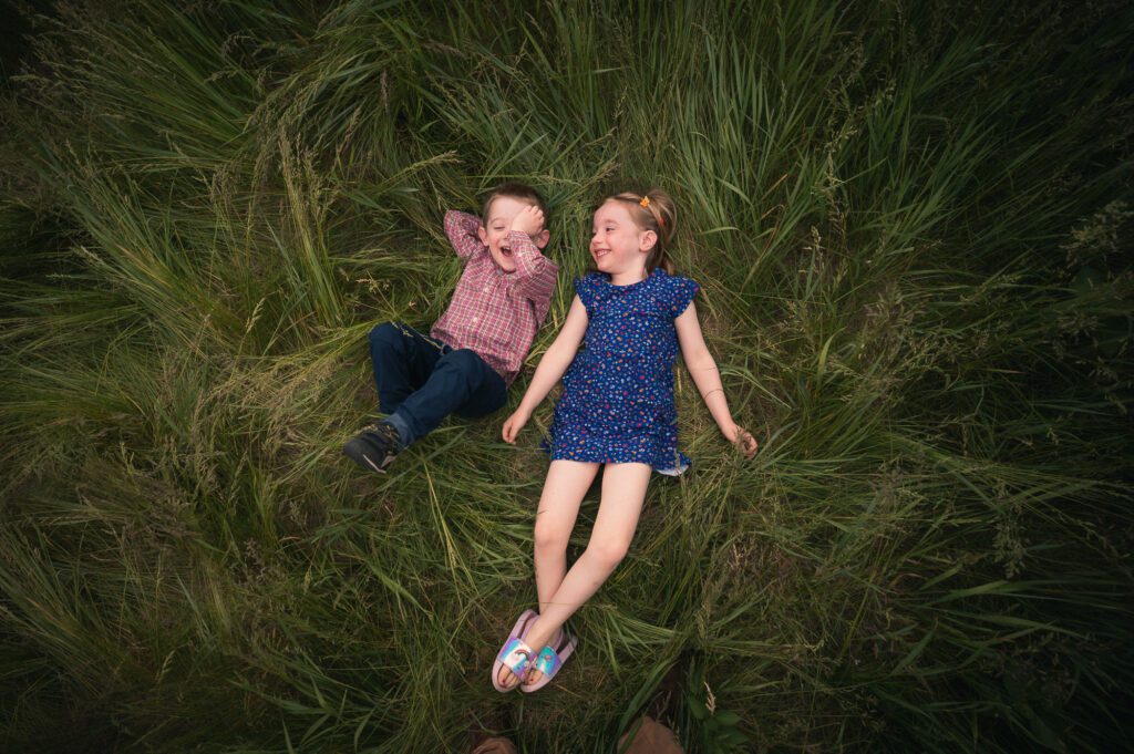 Two children lying in tall green grass, laughing and looking at each other during a relaxed and candid family photo session.