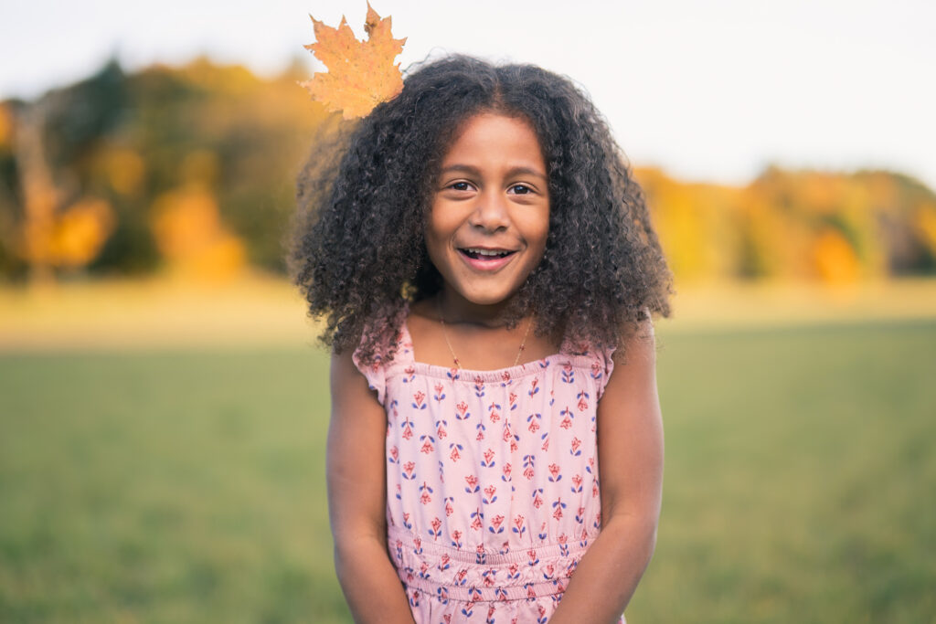 A girl with a maple leaf in her hair smiles in front of bright fall foliage during a family photoshoot outside Boston.