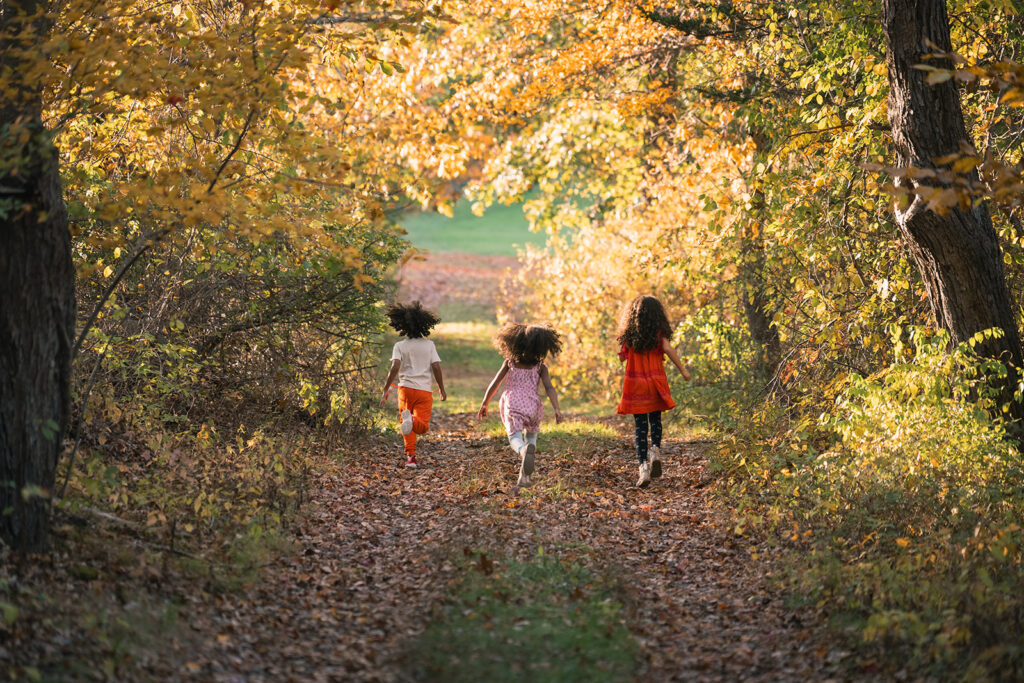 Three children running down a leaf-covered path surrounded by golden autumn trees during a Fall family photo session at a farm in Lincoln, MA by Boston photographer Corey Flint.