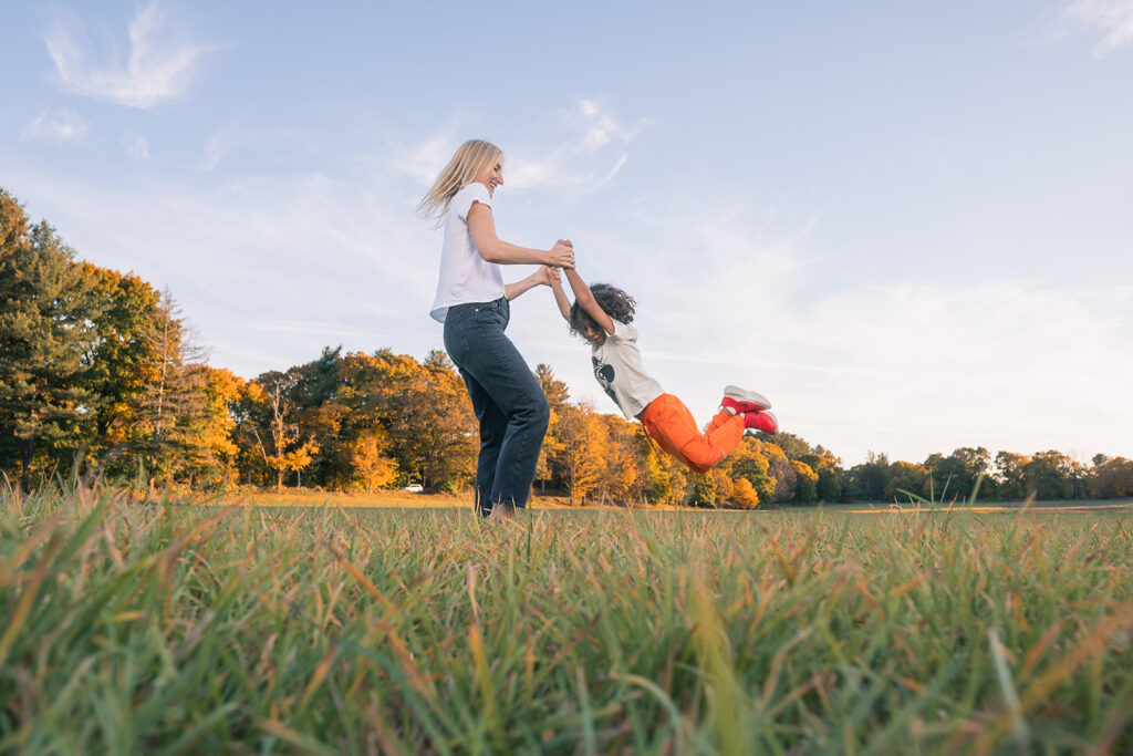 A woman spinning a laughing child through the air in a sunny open field during a Fall family photo session at a farm in Lincoln, MA by Boston photographer Corey Flint.