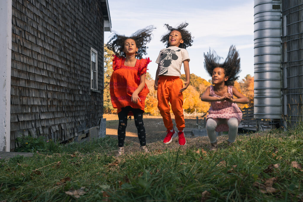 Three kids jumping and laughing between two barn buildings during a Fall family photo session at a farm in Lincoln, MA by Boston photographer Corey Flint.