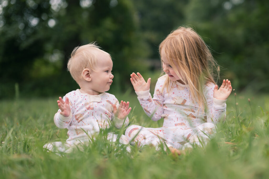 Two young children in matching pajamas sit on the grass, happily playing together and raising their hands in a joyful moment.
