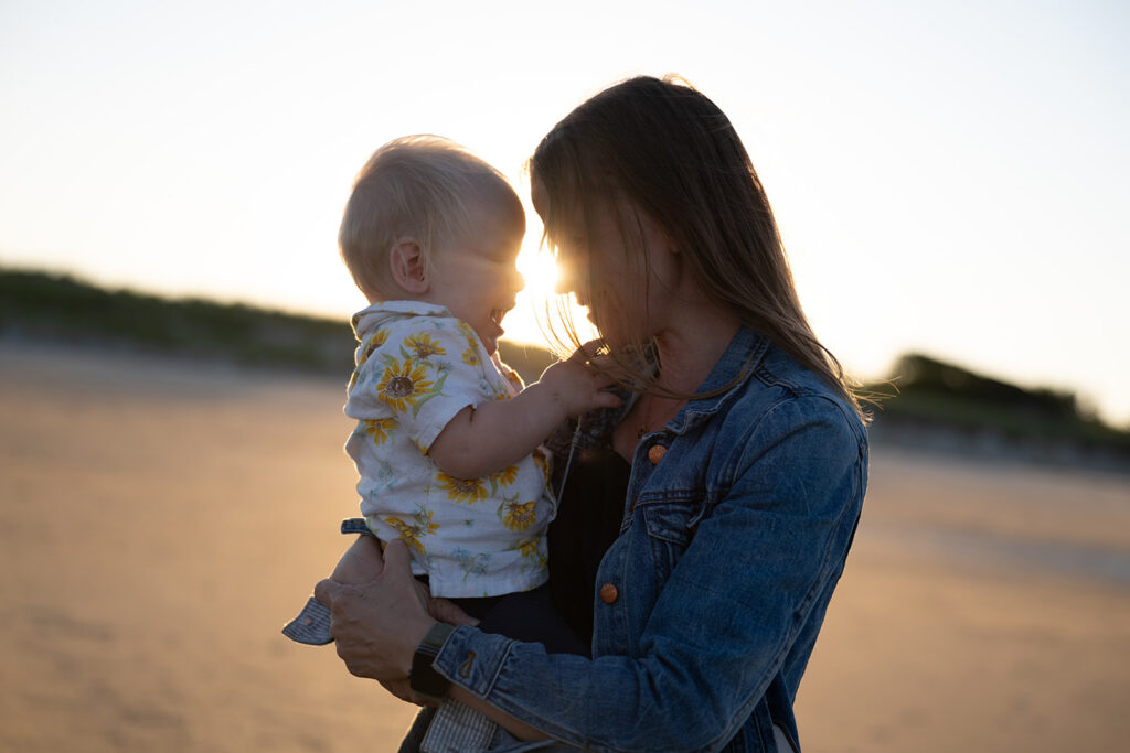 A mother holds her baby in her arms at sunset on the beach, sharing a tender moment as the sun shines through their faces.
