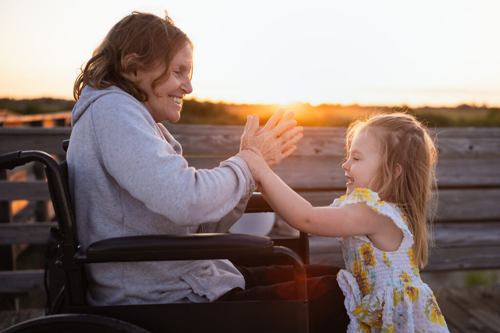 A joyful moment between a grandmother in a wheelchair and her granddaughter, as they play together at sunset, clapping hands and sharing smiles.

