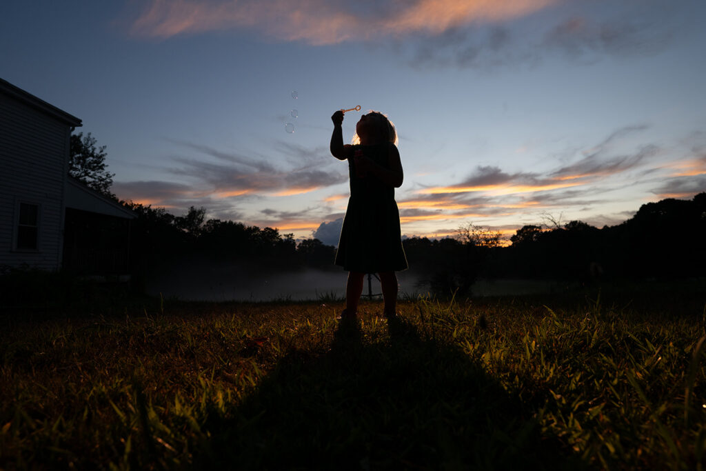 A child blows bubbles at sunset, their silhouette framed against the vibrant sky and tranquil landscape.
