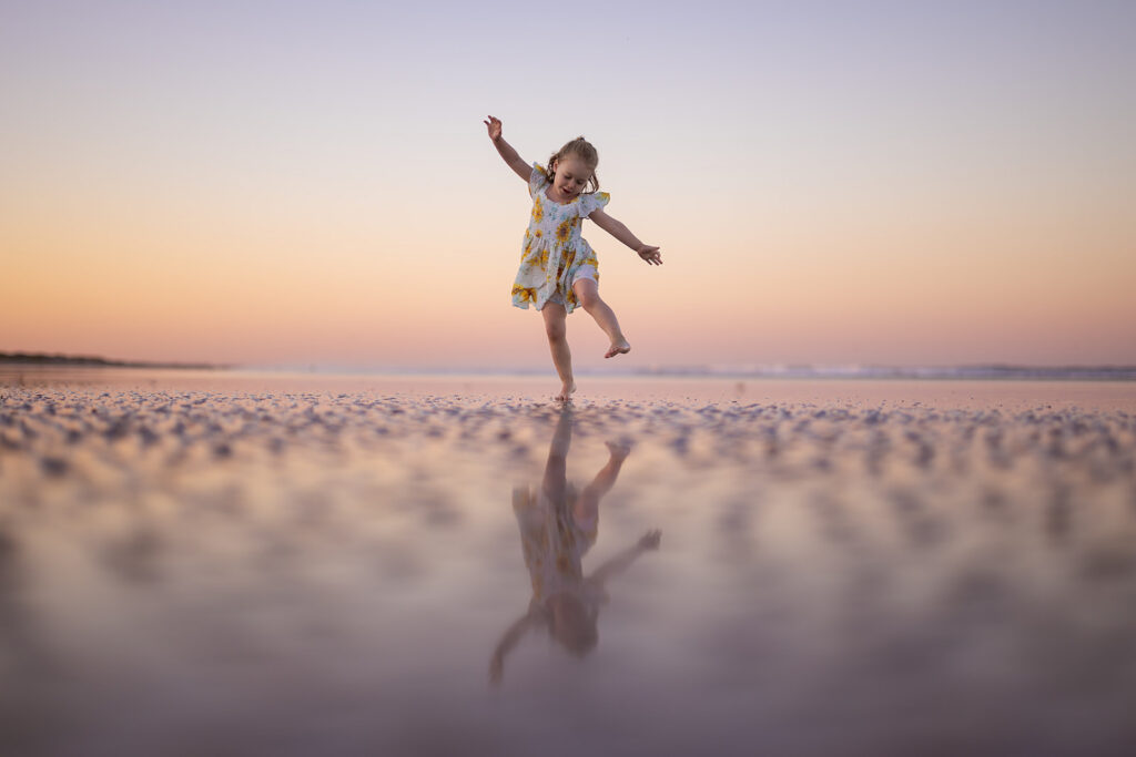 A young girl in a yellow flowered dress joyfully skips along the beach at sunset, her reflection visible in the wet sand.
