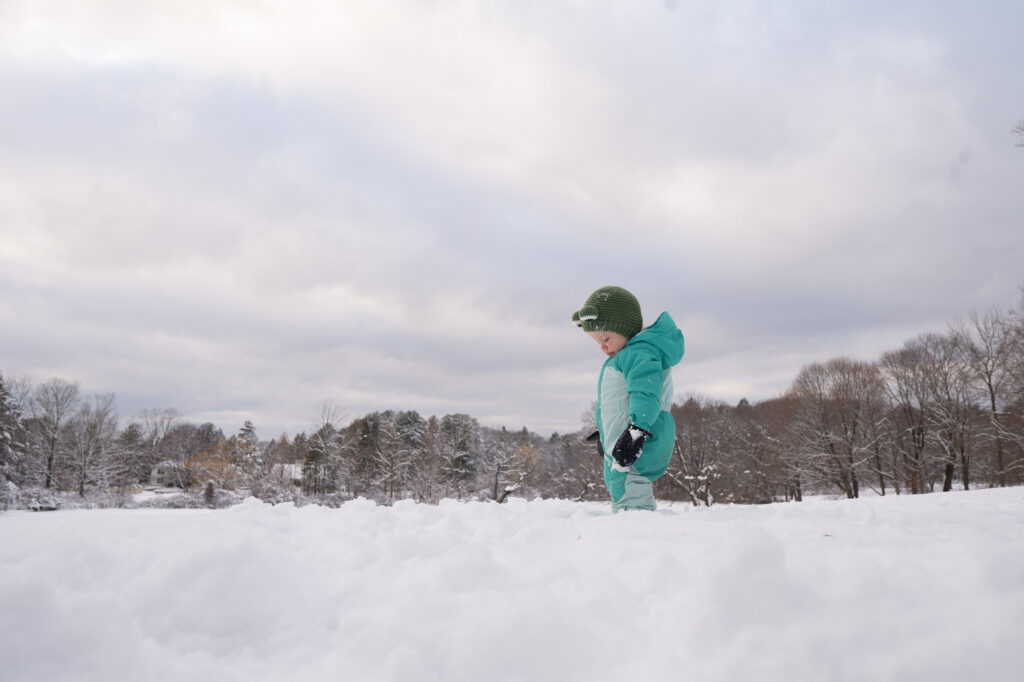Toddler in teal snowsuit standing in snowy open field during Lincoln MA winter family photos - Corey Flint Photography