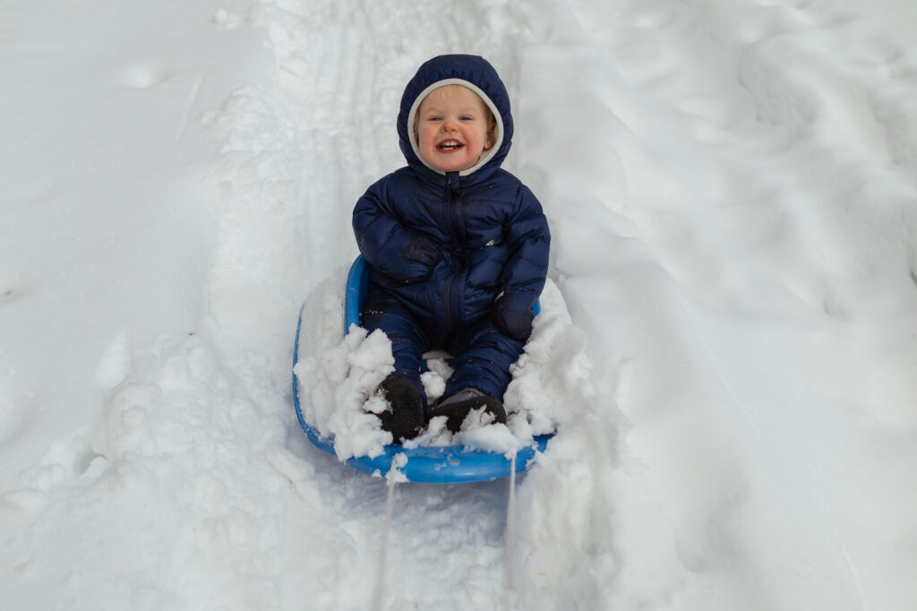 Laughing toddler on blue sled during winter family session with Lincoln Massachusetts family photographer - Corey Flint Photography