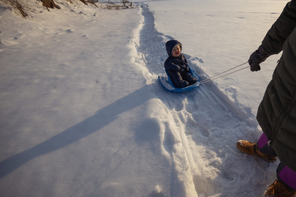 Parent pulling child on sled across snow in golden afternoon light at Lincoln MA winter family session - Corey Flint Photography