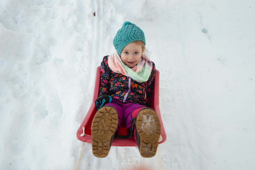 Girl in red sled laughing during MetroWest winter family photography session - Corey Flint Photography