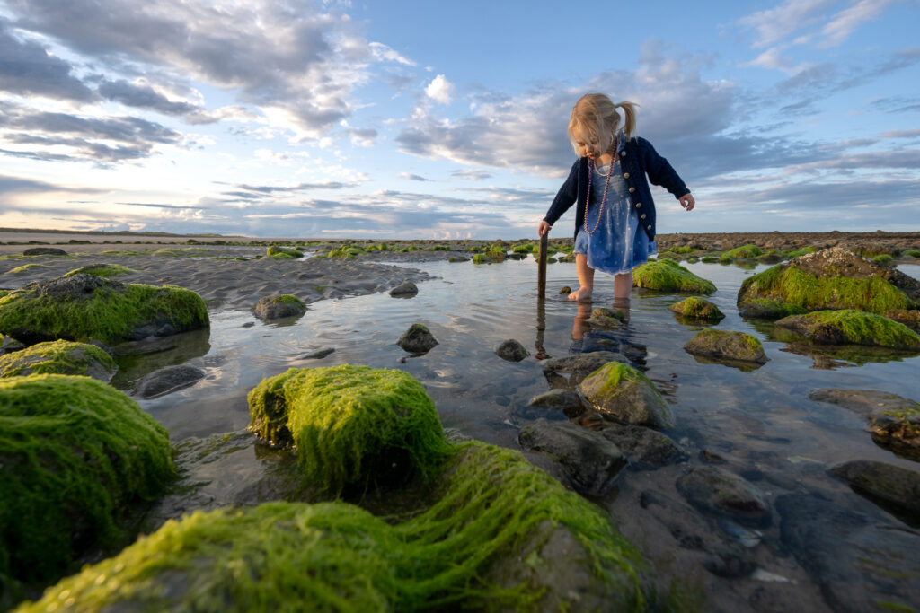 Young girl crouching at a Maine tide pool, photographed from low angle with dramatic coastal sky — award-winning family photography by Corey Flint, Lincoln MA