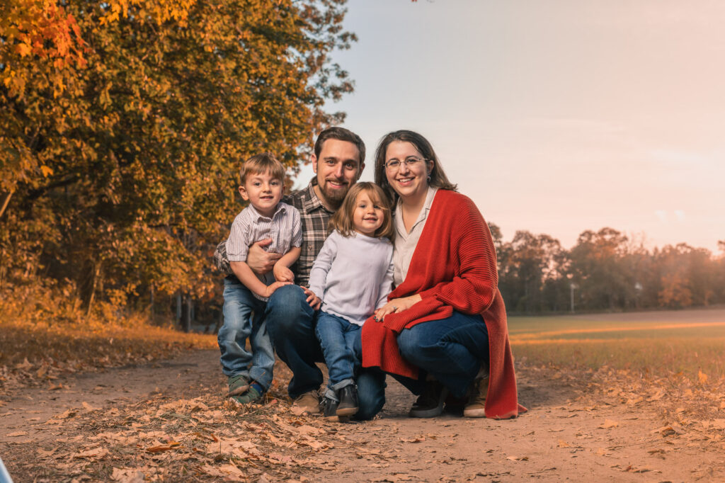 Family of four on a fall dirt path at golden hour, mom in red cardigan and glasses, dad in plaid flannel, kids in soft neutrals, New England family photography