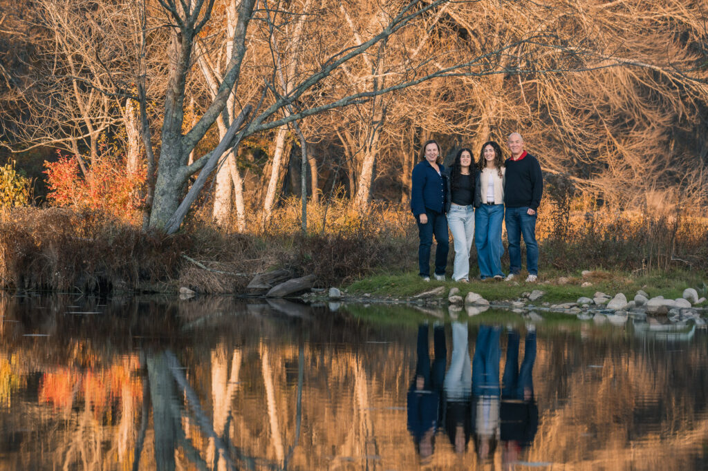 Family of four including two teenagers reflected in a pond, late fall New England foliage
