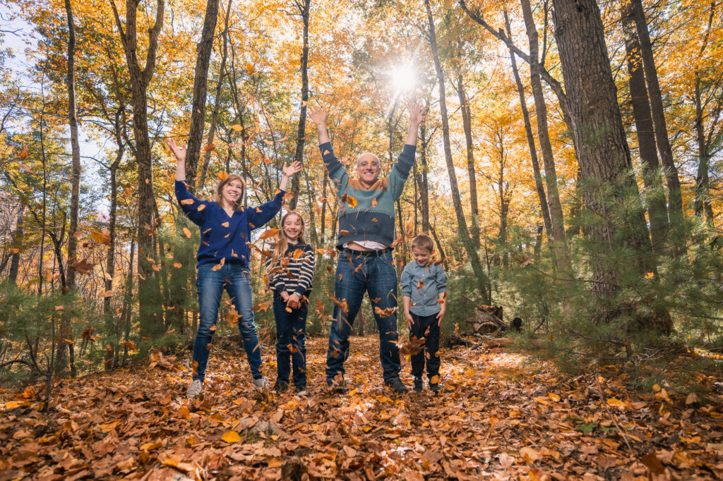 Family of four throwing fall leaves in the air, laughing and jumping, arms raised in New England forest