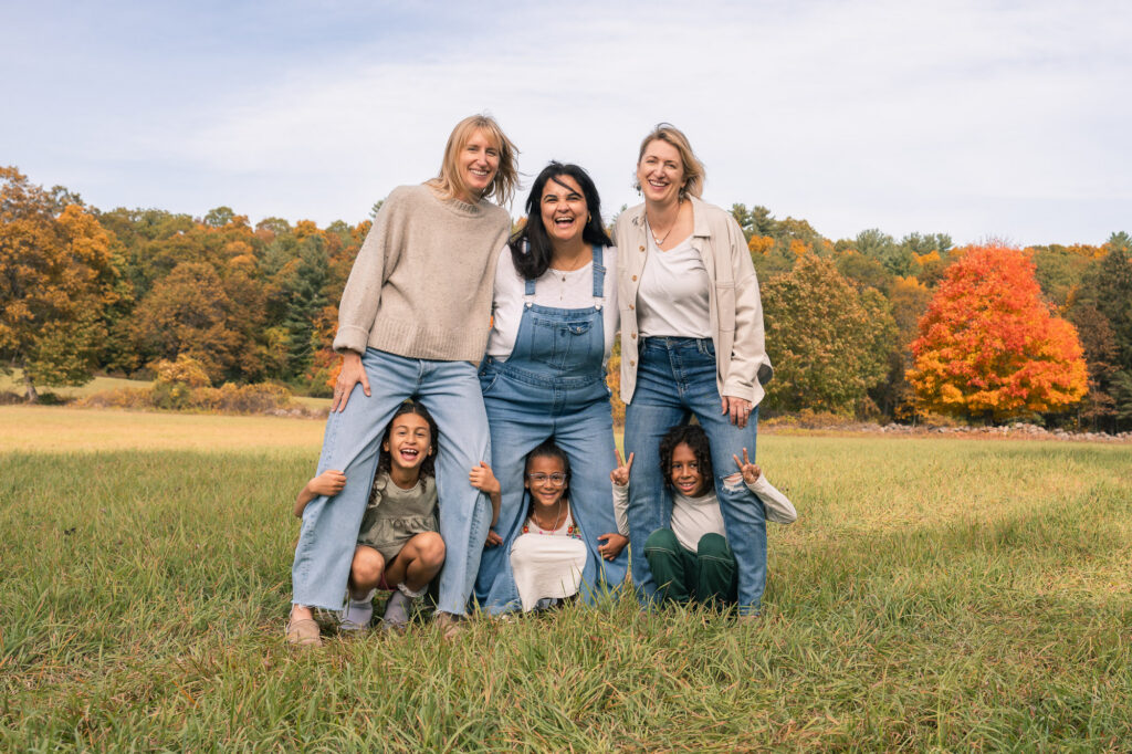 Three moms laughing while kids peek through their legs in a fall field, Massachusetts family photography