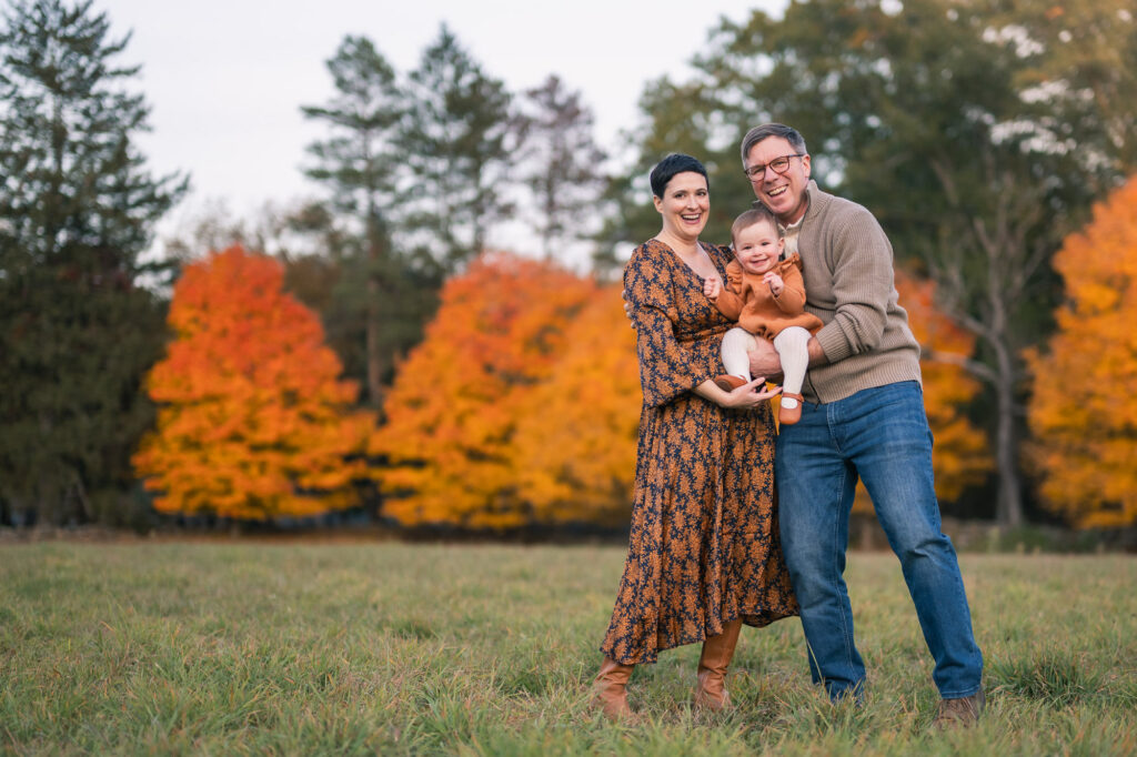 Mom in rust and navy floral dress holding baby, dad in tan sweater and jeans, fall foliage background