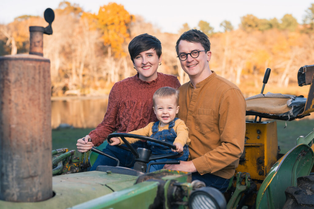 Family of three on a vintage green John Deere tractor at a New England farm, fall colors in background