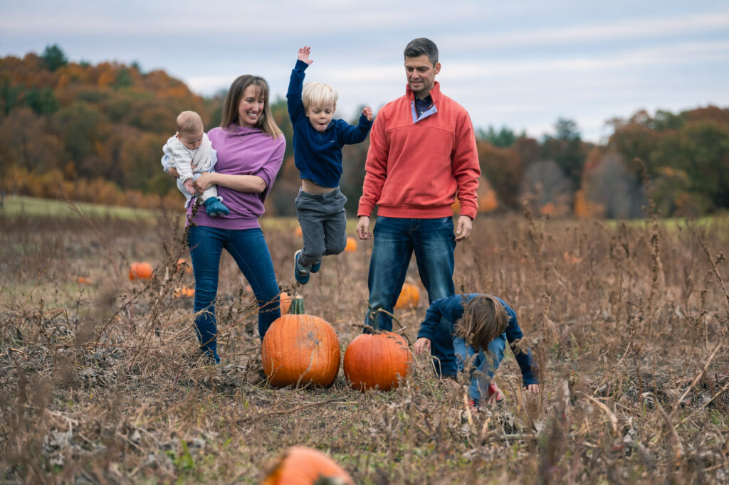 Parents laughing while three kids climb all over them in fall pumpkin field, casual family photos