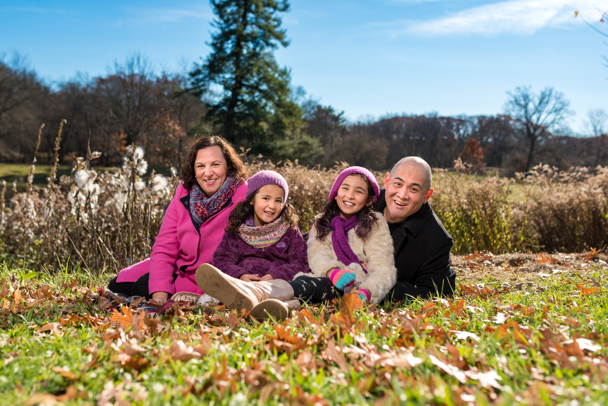 Parents and two young daughters in pink and purple hats sitting among fallen autumn leaves