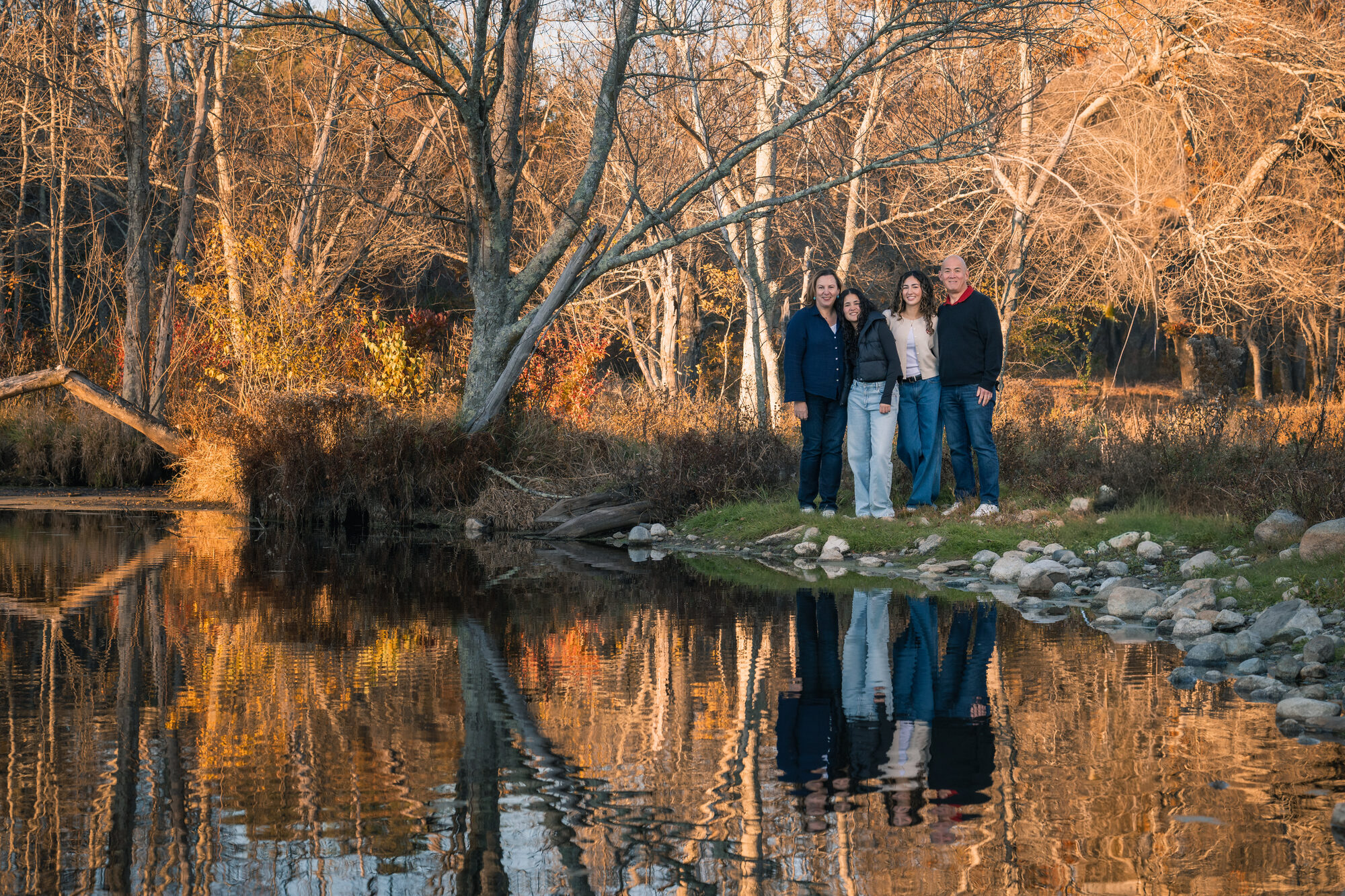 Family of four standing beside a still pond with a mirror reflection at golden hour in Lincoln, MA