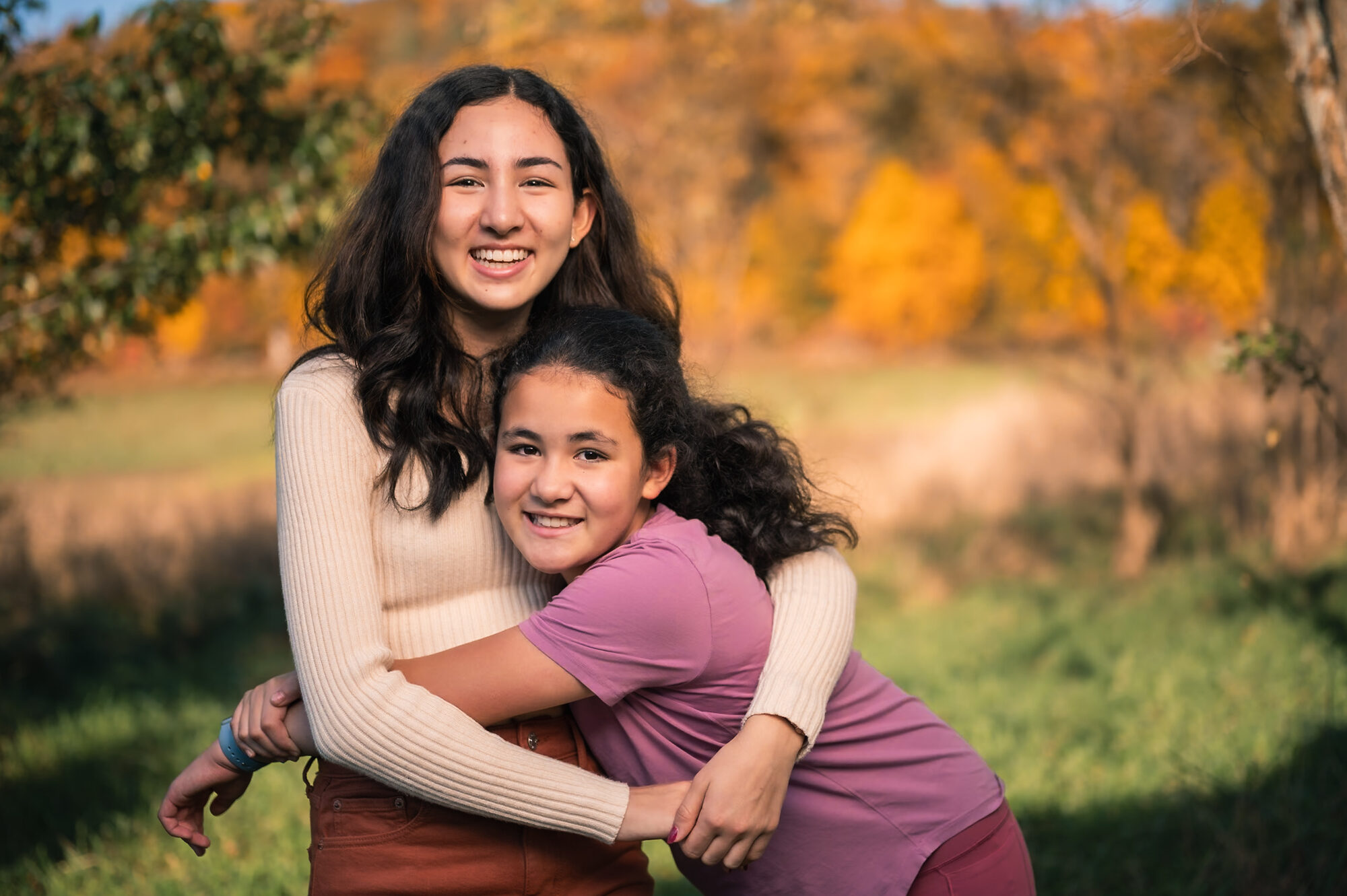 An older and younger sister hugging and laughing in a sunny autumn field in Lincoln, MA