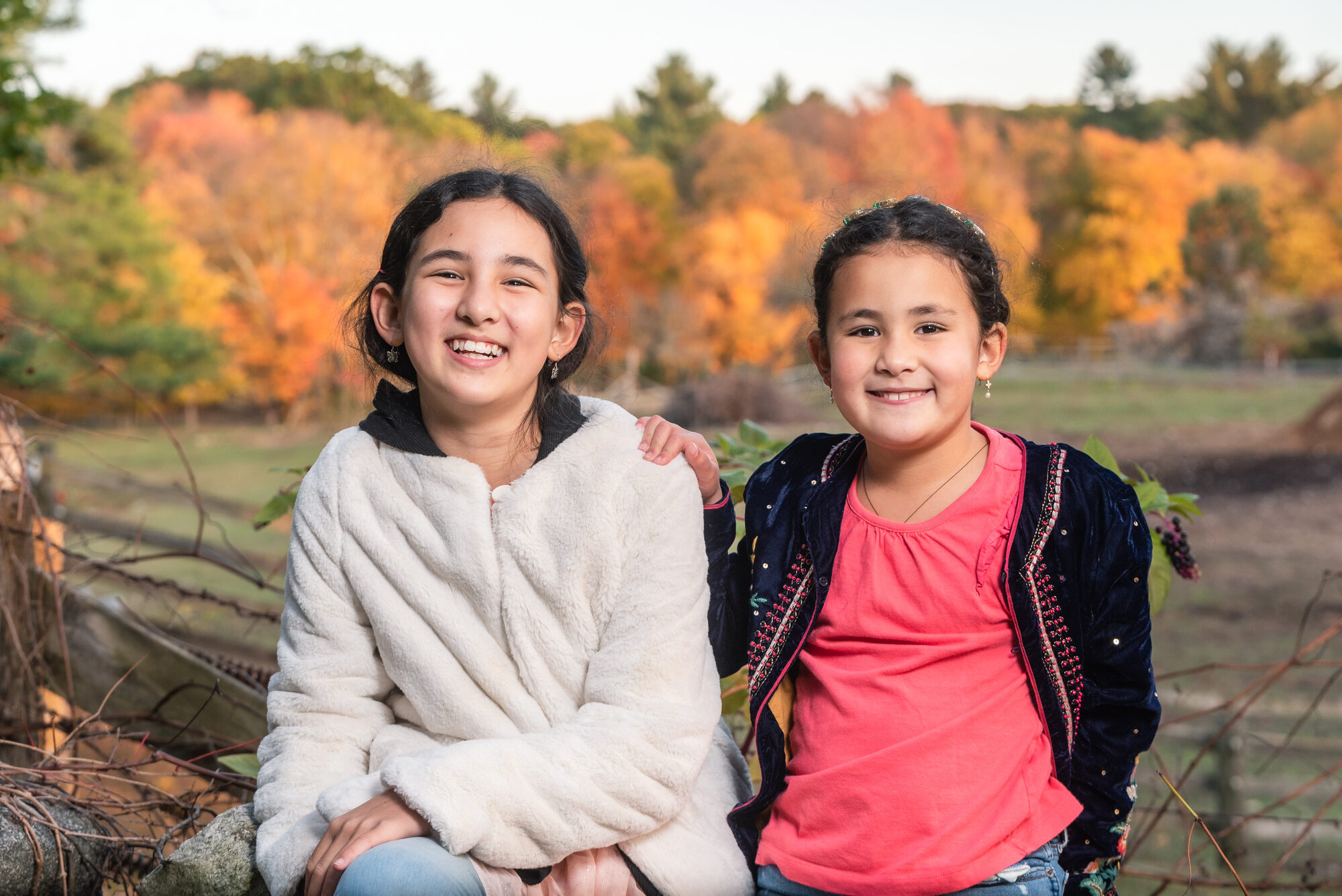 Two young sisters smiling in front of bright orange fall foliage at a Lincoln, MA farm