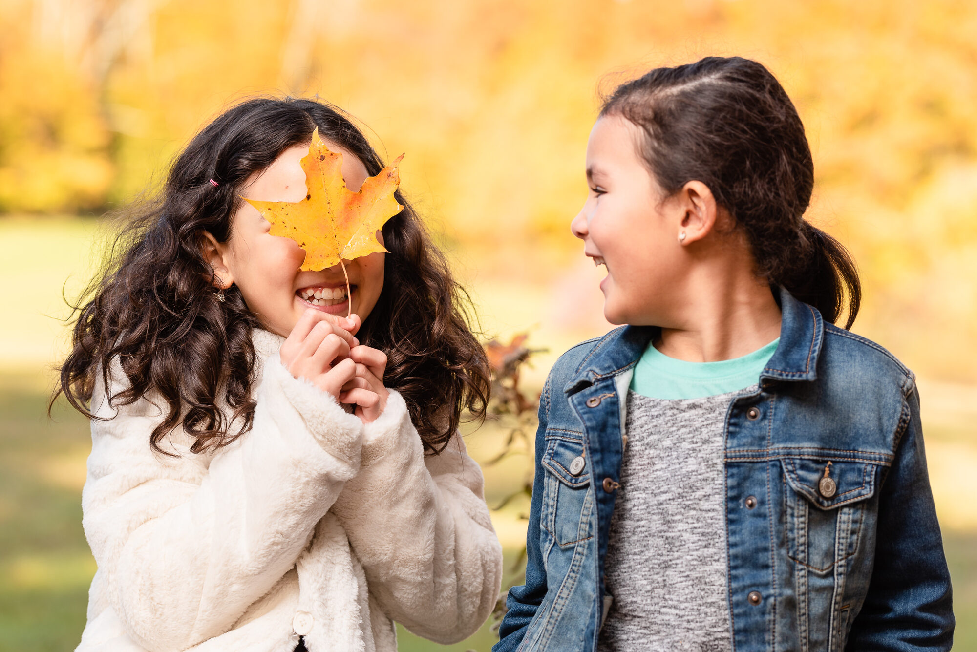 Two young girls laughing, one holding an orange maple leaf in front of her face, during a fall portrait session
