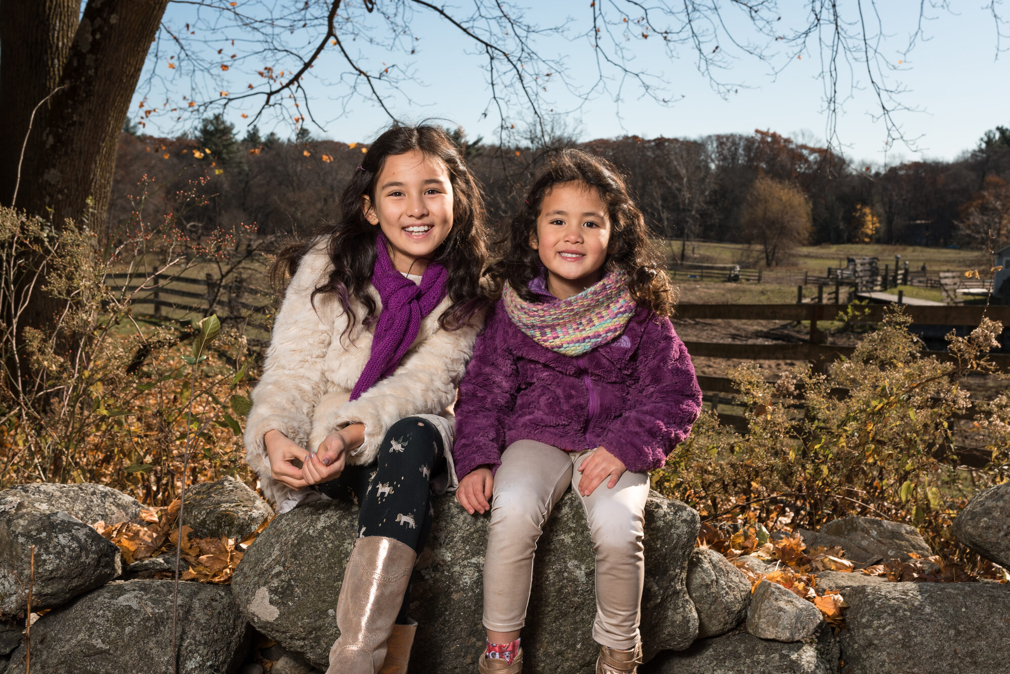 Two young sisters in cozy coats sitting on a stone wall with a farm fence behind them