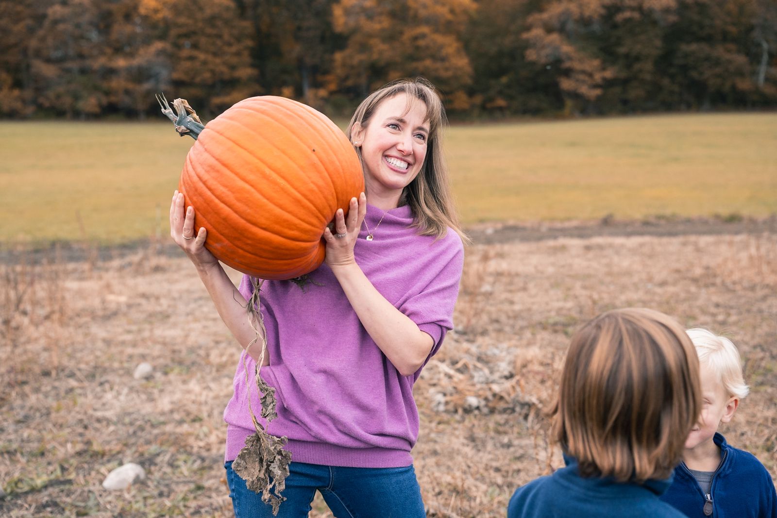 A laughing mom holding a huge pumpkin on her shoulder while her two boys watch at Flint Farm