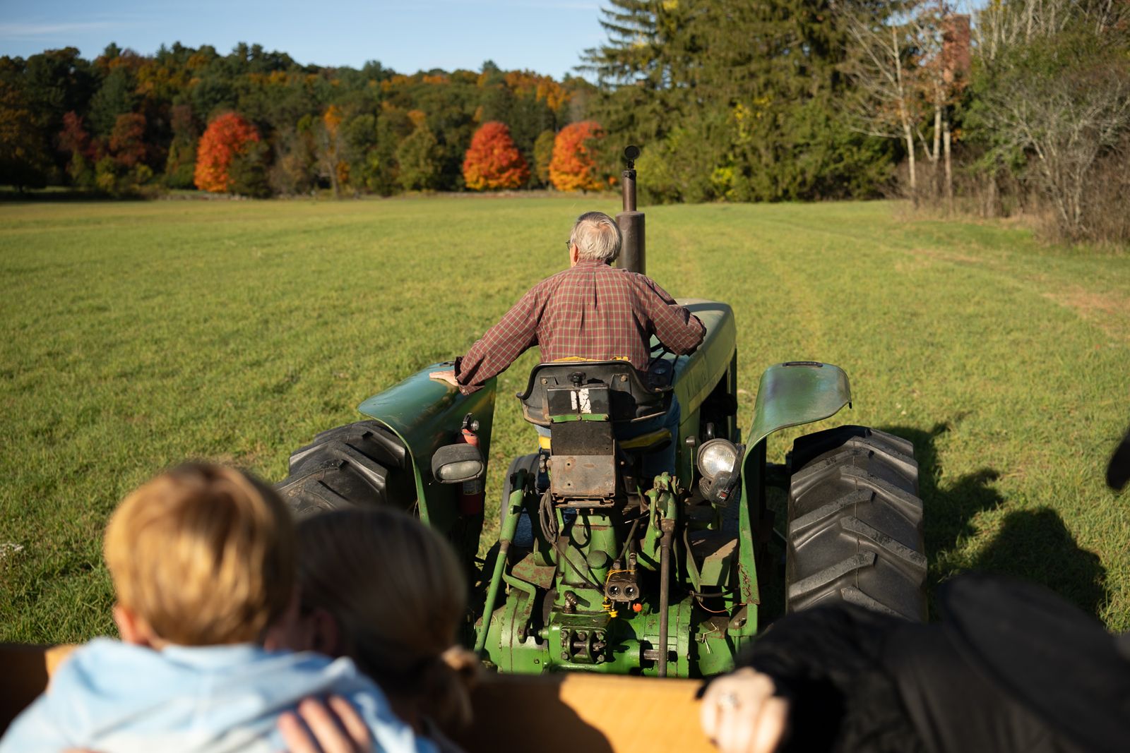 View from the hay wagon behind a green tractor crossing a field lined with red and orange fall foliage in Lincoln, MA